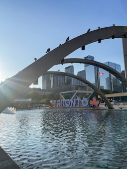 An urban scene featuring a large reflective pool with modern arched bridges. In the background, high-rise buildings create a cityscape, complemented by a Canadian flag fluttering in the wind. The word 'TORONTO' is prominently displayed along the waterfront accompanied by a red maple leaf symbol. The sun casts a bright flare from the left, adding a dynamic element to the composition.
