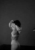 A person wearing underwear is lifting a garment over their head, partially obscuring their face. The image is monochrome, highlighting shadows and contours against a dark background. A tattoo is visible on their left side.