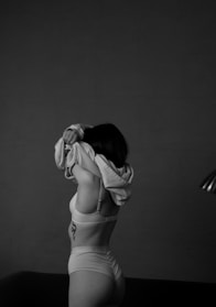 A person wearing underwear is lifting a garment over their head, partially obscuring their face. The image is monochrome, highlighting shadows and contours against a dark background. A tattoo is visible on their left side.