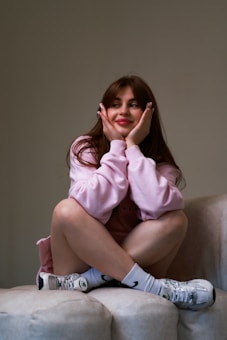 A young woman with long brown hair is sitting on a beige couch. She is wearing a pink sweatshirt and white athletic sneakers with black details. Her expression is thoughtful and content as she rests her chin on her hands, elbows on her knees.