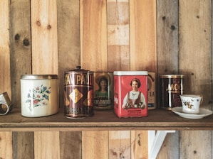 A wooden shelf displays a collection of vintage tins, each with unique designs and illustrations. The tins vary in color and feature intricate graphics, including floral patterns, portraits, and bold text. A small, delicate teacup with a saucer is positioned to the right of the tins.