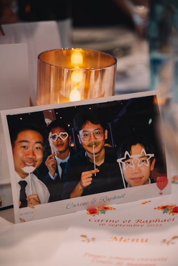 A photo displayed on a table shows four people posing with playful accessories like paper glasses and mustache props in a festive ambiance. The table has a candle and printed materials related to an event, including the names Carine and Rapha&euml;l and the date 10 September 2023.