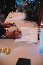 A person is writing on an open book placed on a table surrounded by small, decorative candles. The person is wearing a bracelet and a ring and holding a pen. The scene is softly lit, creating a warm and intimate atmosphere.