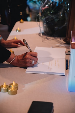 A person is writing on an open book placed on a table surrounded by small, decorative candles. The person is wearing a bracelet and a ring and holding a pen. The scene is softly lit, creating a warm and intimate atmosphere.