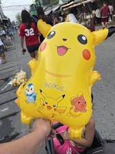 A yellow Pikachu balloon is held by a hand in an outdoor market setting. The balloon features images of Pikachu and other Pokémon characters, including Piplup and a pink Pokémon. People casually walk in the background, including someone in a red shirt with the number 32. The setting appears lively and bustling.