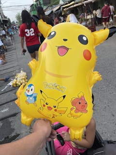A yellow Pikachu balloon is held by a hand in an outdoor market setting. The balloon features images of Pikachu and other Pokémon characters, including Piplup and a pink Pokémon. People casually walk in the background, including someone in a red shirt with the number 32. The setting appears lively and bustling.