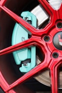 A close-up view of a red alloy wheel with a visible turquoise brake caliper. The design includes intricate spoke patterns and metallic elements. The wheel and brake appear new and polished.