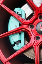 A close-up view of a red alloy wheel with a visible turquoise brake caliper. The design includes intricate spoke patterns and metallic elements. The wheel and brake appear new and polished.
