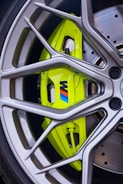 A detailed close-up of a shiny, modern alloy wheel with a prominent lime green brake caliper featuring a logo. The wheel spokes are sleek and metallic, framing the brightly colored caliper and the disc brake behind it. The overall design highlights precision engineering and performance aesthetics.