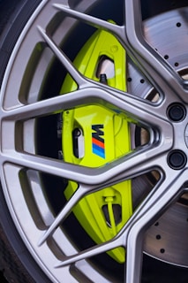 A detailed close-up of a shiny, modern alloy wheel with a prominent lime green brake caliper featuring a logo. The wheel spokes are sleek and metallic, framing the brightly colored caliper and the disc brake behind it. The overall design highlights precision engineering and performance aesthetics.