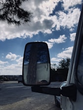 A truck's side mirror reflecting a tree trunk. The scene includes a partly cloudy sky with bright blue tones and scattered white clouds. The view encompasses a road with other vehicles and some distant buildings. Branches from another tree are visible at the top left corner.