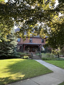 A charming house with a brick facade and a covered porch is surrounded by lush greenery. A tree in the foreground casts dappled shade on the well-maintained lawn and concrete pathway leading to the entrance.