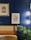 The image features a stylish interior setting with a deep blue paneled wall. Two framed artworks hang on the wall, one with a golden frame depicting a detailed architectural design, and the other a modern black and white abstract leaf design. A modern wall sconce with a double globe light fixture provides illumination. A plush leather or faux leather seating area is visible, along with a natural-looking plant adding a touch of greenery.