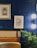 The image features a stylish interior setting with a deep blue paneled wall. Two framed artworks hang on the wall, one with a golden frame depicting a detailed architectural design, and the other a modern black and white abstract leaf design. A modern wall sconce with a double globe light fixture provides illumination. A plush leather or faux leather seating area is visible, along with a natural-looking plant adding a touch of greenery.