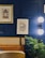 The image features a stylish interior setting with a deep blue paneled wall. Two framed artworks hang on the wall, one with a golden frame depicting a detailed architectural design, and the other a modern black and white abstract leaf design. A modern wall sconce with a double globe light fixture provides illumination. A plush leather or faux leather seating area is visible, along with a natural-looking plant adding a touch of greenery.