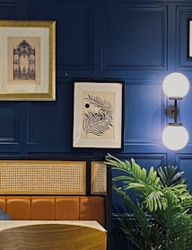 The image features a stylish interior setting with a deep blue paneled wall. Two framed artworks hang on the wall, one with a golden frame depicting a detailed architectural design, and the other a modern black and white abstract leaf design. A modern wall sconce with a double globe light fixture provides illumination. A plush leather or faux leather seating area is visible, along with a natural-looking plant adding a touch of greenery.