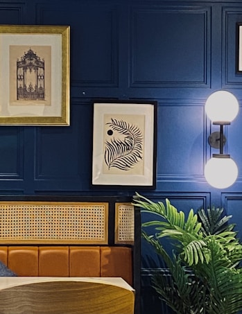 The image features a stylish interior setting with a deep blue paneled wall. Two framed artworks hang on the wall, one with a golden frame depicting a detailed architectural design, and the other a modern black and white abstract leaf design. A modern wall sconce with a double globe light fixture provides illumination. A plush leather or faux leather seating area is visible, along with a natural-looking plant adding a touch of greenery.