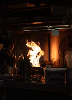 A dimly lit kitchen scene features a large, vigorous flame rising from a stovetop, suggesting intense cooking activity. A person stands nearby, possibly adding ingredients. Various kitchen items, including bottles and plates, are visible on the counter, emphasizing a busy and functional kitchen setting.