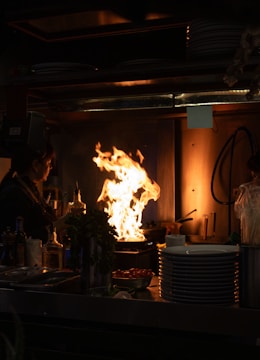 A dimly lit kitchen scene features a large, vigorous flame rising from a stovetop, suggesting intense cooking activity. A person stands nearby, possibly adding ingredients. Various kitchen items, including bottles and plates, are visible on the counter, emphasizing a busy and functional kitchen setting.