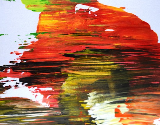 A close-up of an abstract painting featuring bold and dynamic brushstrokes. The colors blend and streak across the canvas, creating a sense of movement and energy. Hues of red, orange, yellow, green, and black dominate the image, with occasional splashes of white.