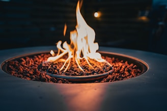A close-up view of a fire pit with flames rising from lava rocks, surrounded by a circular metal structure. The warm glow of the fire contrasts against the dark background, creating a cozy and inviting atmosphere.
