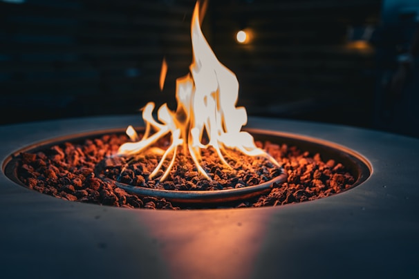 A close-up view of a fire pit with flames rising from lava rocks, surrounded by a circular metal structure. The warm glow of the fire contrasts against the dark background, creating a cozy and inviting atmosphere.
