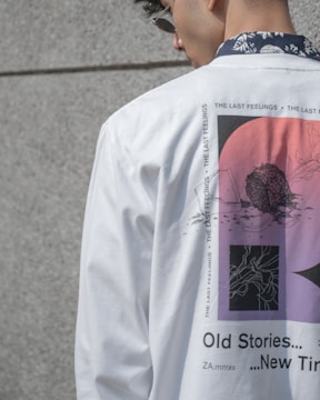 A person is wearing a white shirt featuring abstract designs with red and black elements and the text 'Old Stories... New Times.' The shirt also includes repetitive text 'THE LAST FEELINGS' along the design. The person is wearing sunglasses and has a floral pattern on the collar of a shirt visible underneath.