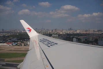 The image depicts a view from an airplane window, showcasing the aircraft's wing with a logo. Below, a sprawling city skyline stretches across the horizon under a partly cloudy sky. The landscape includes numerous high-rise buildings, a mix of green areas, and urban developments.