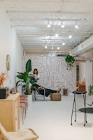 A modern, minimalist interior with bright white walls and a high ceiling featuring exposed beams and lighting fixtures. The space includes a stylish wall with abstract patterns and an assortment of indoor plants. A person is working with another person seated on a chair in the middle of the room, suggesting a relaxed, professional environment. Additional seating with pink chairs and a wooden cabinet are visible to the side.