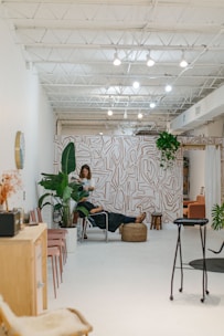 A modern, minimalist interior with bright white walls and a high ceiling featuring exposed beams and lighting fixtures. The space includes a stylish wall with abstract patterns and an assortment of indoor plants. A person is working with another person seated on a chair in the middle of the room, suggesting a relaxed, professional environment. Additional seating with pink chairs and a wooden cabinet are visible to the side.