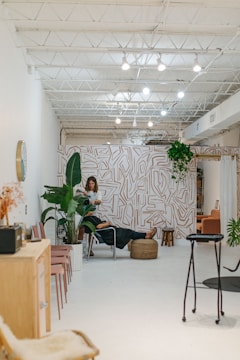 A modern, minimalist interior with bright white walls and a high ceiling featuring exposed beams and lighting fixtures. The space includes a stylish wall with abstract patterns and an assortment of indoor plants. A person is working with another person seated on a chair in the middle of the room, suggesting a relaxed, professional environment. Additional seating with pink chairs and a wooden cabinet are visible to the side.