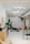 A modern, minimalist interior with bright white walls and a high ceiling featuring exposed beams and lighting fixtures. The space includes a stylish wall with abstract patterns and an assortment of indoor plants. A person is working with another person seated on a chair in the middle of the room, suggesting a relaxed, professional environment. Additional seating with pink chairs and a wooden cabinet are visible to the side.