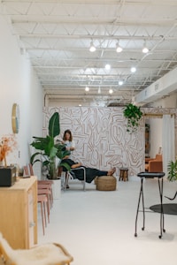 A modern, minimalist interior with bright white walls and a high ceiling featuring exposed beams and lighting fixtures. The space includes a stylish wall with abstract patterns and an assortment of indoor plants. A person is working with another person seated on a chair in the middle of the room, suggesting a relaxed, professional environment. Additional seating with pink chairs and a wooden cabinet are visible to the side.