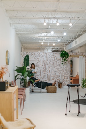 A modern, minimalist interior with bright white walls and a high ceiling featuring exposed beams and lighting fixtures. The space includes a stylish wall with abstract patterns and an assortment of indoor plants. A person is working with another person seated on a chair in the middle of the room, suggesting a relaxed, professional environment. Additional seating with pink chairs and a wooden cabinet are visible to the side.