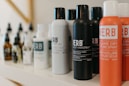 A variety of haircare products are displayed on a shelf. The prominently visible bottles feature different types of sprays and oils with bold labels. The products are arranged in a neat line, showcasing their branding and color schemes.