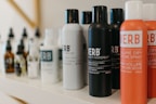 A variety of haircare products are displayed on a shelf. The prominently visible bottles feature different types of sprays and oils with bold labels. The products are arranged in a neat line, showcasing their branding and color schemes.
