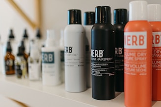A variety of haircare products are displayed on a shelf. The prominently visible bottles feature different types of sprays and oils with bold labels. The products are arranged in a neat line, showcasing their branding and color schemes.