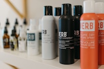 A variety of haircare products are displayed on a shelf. The prominently visible bottles feature different types of sprays and oils with bold labels. The products are arranged in a neat line, showcasing their branding and color schemes.