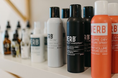 A variety of haircare products are displayed on a shelf. The prominently visible bottles feature different types of sprays and oils with bold labels. The products are arranged in a neat line, showcasing their branding and color schemes.