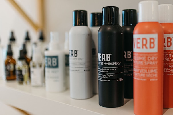 A variety of haircare products are displayed on a shelf. The prominently visible bottles feature different types of sprays and oils with bold labels. The products are arranged in a neat line, showcasing their branding and color schemes.