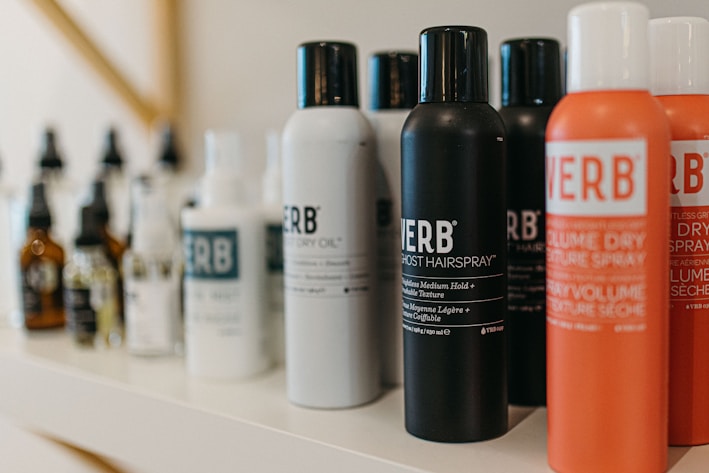 A variety of haircare products are displayed on a shelf. The prominently visible bottles feature different types of sprays and oils with bold labels. The products are arranged in a neat line, showcasing their branding and color schemes.