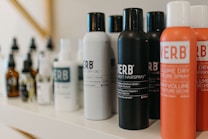 A variety of haircare products are displayed on a shelf. The prominently visible bottles feature different types of sprays and oils with bold labels. The products are arranged in a neat line, showcasing their branding and color schemes.