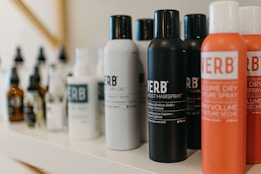 A variety of haircare products are displayed on a shelf. The prominently visible bottles feature different types of sprays and oils with bold labels. The products are arranged in a neat line, showcasing their branding and color schemes.