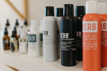 A variety of haircare products are displayed on a shelf. The prominently visible bottles feature different types of sprays and oils with bold labels. The products are arranged in a neat line, showcasing their branding and color schemes.