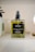 A rectangular glass bottle of body oil labeled 'little barn apothecary HONEYSUCKLE + GRAPEFRUIT BODY OIL' with a black pump on top. The bottle contains a yellow-green liquid and is placed on a shelf with a blurred background.