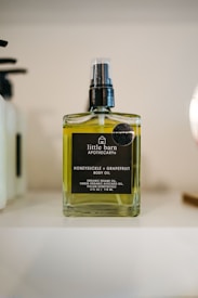 A rectangular glass bottle of body oil labeled 'little barn apothecary HONEYSUCKLE + GRAPEFRUIT BODY OIL' with a black pump on top. The bottle contains a yellow-green liquid and is placed on a shelf with a blurred background.