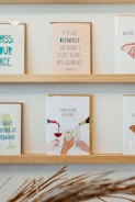 On a wooden shelf, various greeting cards are displayed. The central card features an illustration of hands clinking glasses, with the text 'Cheers to pour decisions.' The other cards include various humorous messages, some referencing life events such as weddings and congratulatory notes.