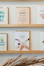 On a wooden shelf, various greeting cards are displayed. The central card features an illustration of hands clinking glasses, with the text 'Cheers to pour decisions.' The other cards include various humorous messages, some referencing life events such as weddings and congratulatory notes.