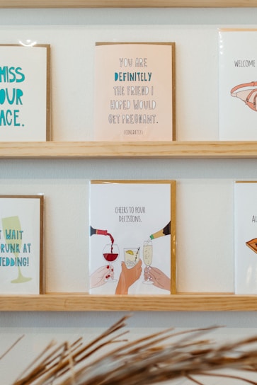 On a wooden shelf, various greeting cards are displayed. The central card features an illustration of hands clinking glasses, with the text 'Cheers to pour decisions.' The other cards include various humorous messages, some referencing life events such as weddings and congratulatory notes.