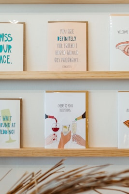 On a wooden shelf, various greeting cards are displayed. The central card features an illustration of hands clinking glasses, with the text 'Cheers to pour decisions.' The other cards include various humorous messages, some referencing life events such as weddings and congratulatory notes.